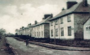 A out of focus black and white photo of a street of houses. A cyclist is moving down the middle of the road in front of the houses.