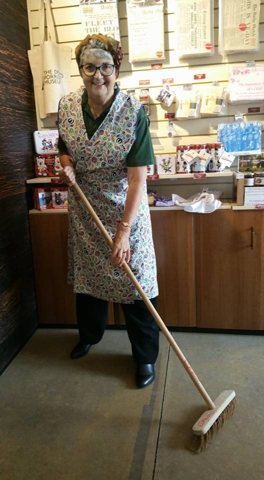 A smiling person dressed in a apron with a broom.