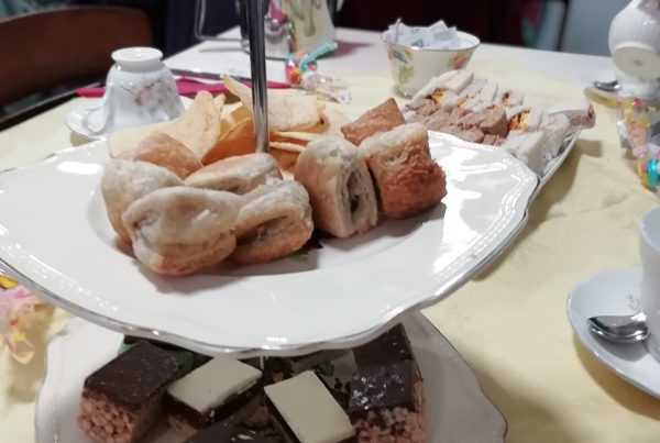 A cake stand with mini sausage rolls, traybakes and crisps on a table. There is also some small sandwiches and teacups on the table. In the background some retirees are sat around a table.