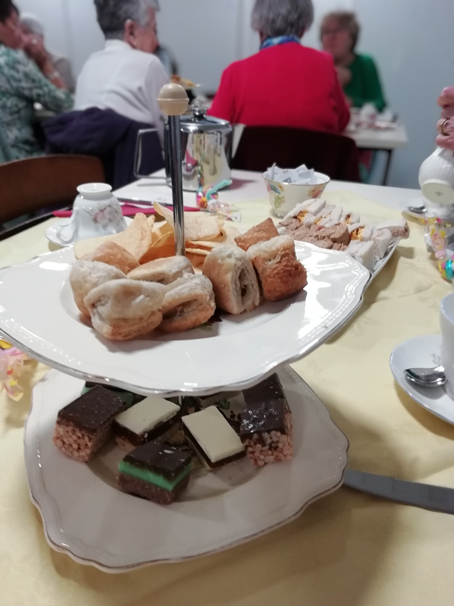 A cake stand with mini sausage rolls, traybakes and crisps on a table. There is also some small sandwiches and teacups on the table. In the background some retirees are sat around a table.