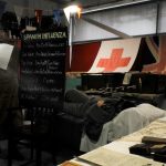 A living history reenactor holds up a blackboard and talks. Another is on a bed behind her.