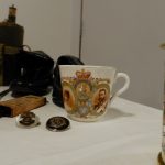 On a table are a selection of objects relating to WW1 such as a cup, some badges and magazines.
