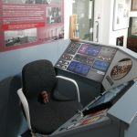 A control desk with a touchscreen and some switches on. There is a chair behind it with a small toy bear on.