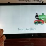 A wee toy bear in front of a digital touchscreen. The touchscreen has a green train on it.