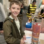 A boy stands next to The Devil's Porridge Museum's time capsule he is holding a letter. Someone dressed as a bee is in the background.