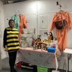 A person wearing a bee costume stands next to a table filled with a display of honey, toys, cushions and other bee related products.