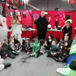 Children in assorted Halloween costumes sit in a circle on the floor. A lady dressed as a witch stands behind them and is talking to them. A lady with green hair sits to the right of the witch and a lady in a white dress stands to the right.