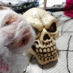 A wee toy bear wearing a spiderweb Halloween costume poses before a glowering plastic skull on the table. The plastic skull is a Halloween decoration.