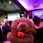A toy bear looking at the camera with people sat at tables behind him. A projection reads "Celebrating Thriving Communities." The Devil's Porridge Museum won this award at the South of Scotland regional final.