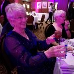 A museum trustee looking happy and sparkly sat at a table. She is holding a toy bear and a drink.