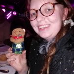A happy person sits at a table and holds a toy bear wearing a kilt.