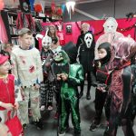 A group of children stood up wearing an assorted jumble of Halloween costumes. Many of them wear Halloween masks. In the background is some Halloween decorations.