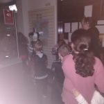 Photo taken in the dark inside the museum, near the entrance. Children in a jumbled assortment of Halloween costumes walk past the front desk in the line.