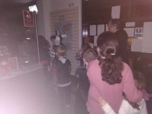 Photo taken in the dark inside the museum, near the entrance. Children in a jumbled assortment of Halloween costumes walk past the front desk in the line.