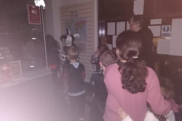 Halloween Party 2023 30 Photo taken in the dark inside the museum, near the entrance. Children in a jumbled assortment of Halloween costumes walk past the front desk in the line.