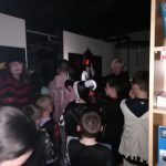 A photo taken in the dark downstairs in The Devil's Porridge Museum. An assortment of children in various Halloween costumes stand with their backs the camera. A lady with a Halloween lantern stands in front of them.