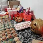 A table adorned with Halloween food, including cupcakes and marshmallows that look like spiders. There are three pumpkin trick or treat bowls filled with crisps, some paper cups, a gold glittery pumpkin and a spiderweb table cloth decorating the table.