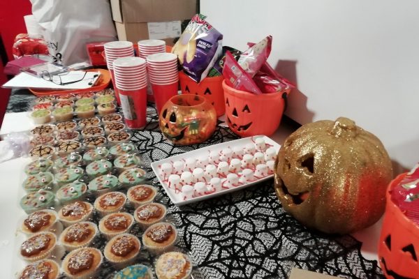 Halloween Party 2023 32 A table adorned with Halloween food, including cupcakes and marshmallows that look like spiders. There are three pumpkin trick or treat bowls filled with crisps, some paper cups, a gold glittery pumpkin and a spiderweb table cloth decorating the table.