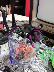 A photo of a glass containing a mystery number of plastic spiders. On top of the glass is a toy spider with googly eyes. In the background part of a Halloween lantern and a toy Halloween TV decoration.