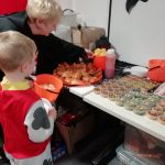 A child dressed as a Dalmatian stands in front of a table containing Halloween themed food in this photo. This includes cupcakes with spiderweb and scary face designs. There is also some hot dogs on the table.