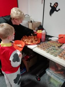 A child dressed as a Dalmatian stands in front of a table containing Halloween themed food in this photo. This includes cupcakes with spiderweb and scary face designs. There is also some hot dogs on the table.