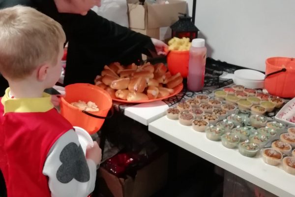 Halloween Party 2023 5 A child dressed as a Dalmatian stands in front of a table containing Halloween themed food in this photo. This includes cupcakes with spiderweb and scary face designs. There is also some hot dogs on the table.