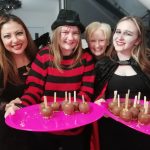 Four happy members of our museum team wearing Halloween costumes. Two of them, dressed as Freddy Krueger and a vampire, hold pink plates full of chocolate apples.