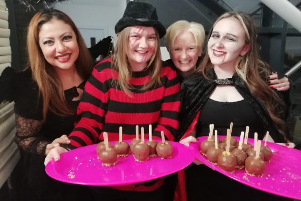 Halloween Party 2023 4 Four happy members of our museum team wearing Halloween costumes. Two of them, dressed as Freddy Krueger and a vampire, hold pink plates full of chocolate apples.