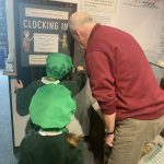 Two school children using one of the museum's clocking in card machines. A museum volunteer stands beside them.