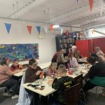 Parents and children are sat at two tables making Halloween crafts together.