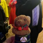 A toy bear wearing a kilt and a bow tie. There are some people in formal attire in the background.