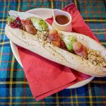 A festive baguette filled with pigs in blankets, Brussel sprouts, stuffing and cranberry sauce. A wee pot of gravy stands on the plate beside the baguette. The logo for The Devil's Porridge Museum's café is in the top left corner.