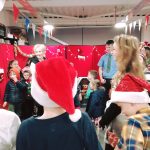 A photo taken behind three children wearing Santa hats. Some more children, festive decorations and members of our museum team can be seen in front of them.