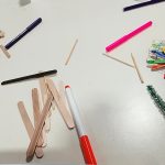 Some felt tip pens, popsicle sticks and pipe cleaners strewn across a table. There is a small Christmas tree and snowflake decoration made out of popsicle sticks on the table.