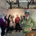 A smiling person is dressed as a munition worker and is stood next a nitrating pan. Some young people and a member of our museum tear are in the background.