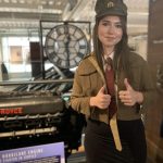 A young lady dressed in part of a replica soldier uniform poses for a photo. The Mossband clock can be seen behind her.