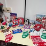 An arrangement of tombola prizes on the table. These include Christmas stockings, a puzzle book, some blue ribbon chocolates, festive tablecloths, a picture frame, a Christmassy puzzle and more.