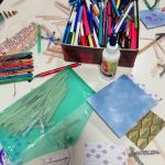 A table full of crafting materials including a tin of felt tip pens, various pieces of paper, popsicle sticks, glue and glittery pipe cleaners.