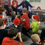 Children sit on the floor in a room decorated for Christmas. They are looking at an adult who is talking to them.