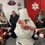 An adult wrapped in paper roll and tinsel by the children at the party. They hold some Christmas baubles on their fingers and a carrot in their mouth, so they are dressed up as a snowman.