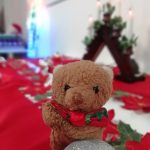 A small toy bear wearing a bow tie with some Christmas decorations in the background.