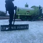 A statue, which reads "Lest we forget" in the snow with Sir James the fireless locomotive in the background. Snow is continuing to fall in the photo.