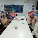 People sat at a table, many of them wear paper Christmas hats. They are clapping their hands and look happy.