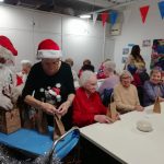 Santa and one of the Museum's Trustees attend a trolly of Christmas gift bags. Some people sit at two rows of tables behind them.