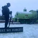 A statue, which reads "Lest we forget" in the snow with Sir James the fireless locomotive in the background. Snow is continuing to fall in the photo.