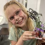 A happy child holds a pot glass pot with a plant in it.