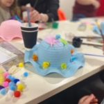 An Easter bonnet amongst craft supplies on a table with a child in the background.
