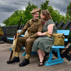 Two living history re-enactors sat on a bench, looking at each other. They are dressed as a soldier and a lady in 1940s clothing.