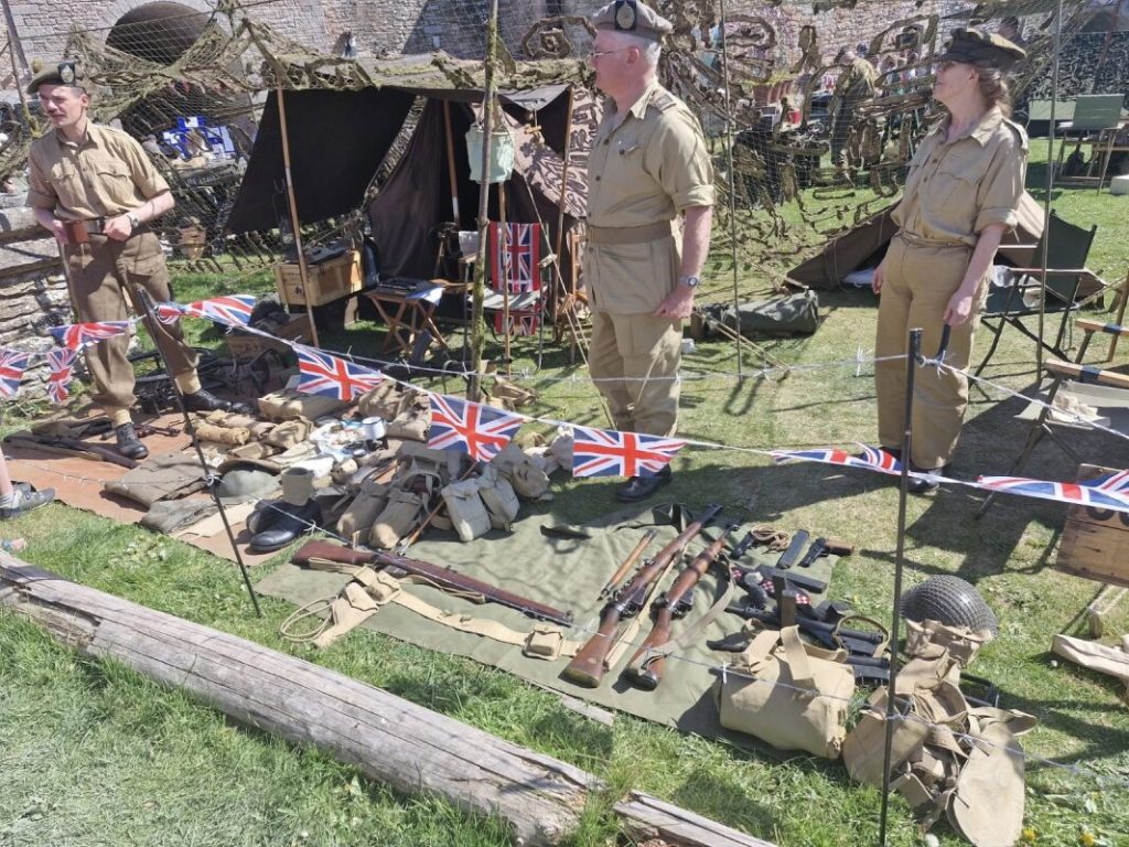 Three living history re-enactors dressed as soldiers with a display of World War Two military items arranged on the ground.