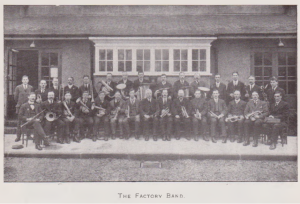 Group of people holding musical instruments sitting in front of a building. This is the Factory Band.